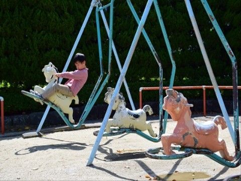 公園にある遊具。鉄製の棒が組まれているところに、馬の乗り物が３つ並んで吊るされている。男の子が１人乗っている。