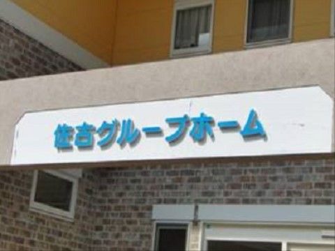 手前に横長のコンクリート造りの看板があり、施設名が水色で表記されている。壁の上部は黄色、下部はレンガ調である。