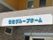 手前に横長のコンクリート造りの看板があり、施設名が水色で表記されている。壁の上部は黄色、下部はレンガ調である。