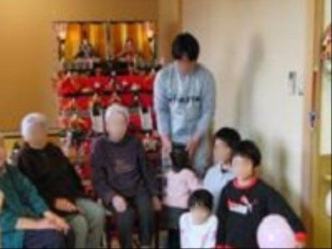 部屋の奥に７段の雛飾りが飾られている。その手前にスタッフや入居者様が集合している。子供の姿も見える。