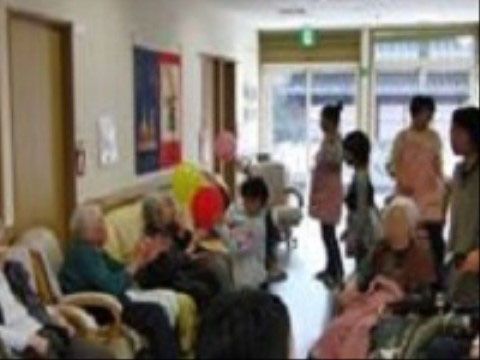 施設の一室に大勢の人が写っている。車椅子や椅子に入居者様たちが座っていて、子供たちが数人一緒にいる。