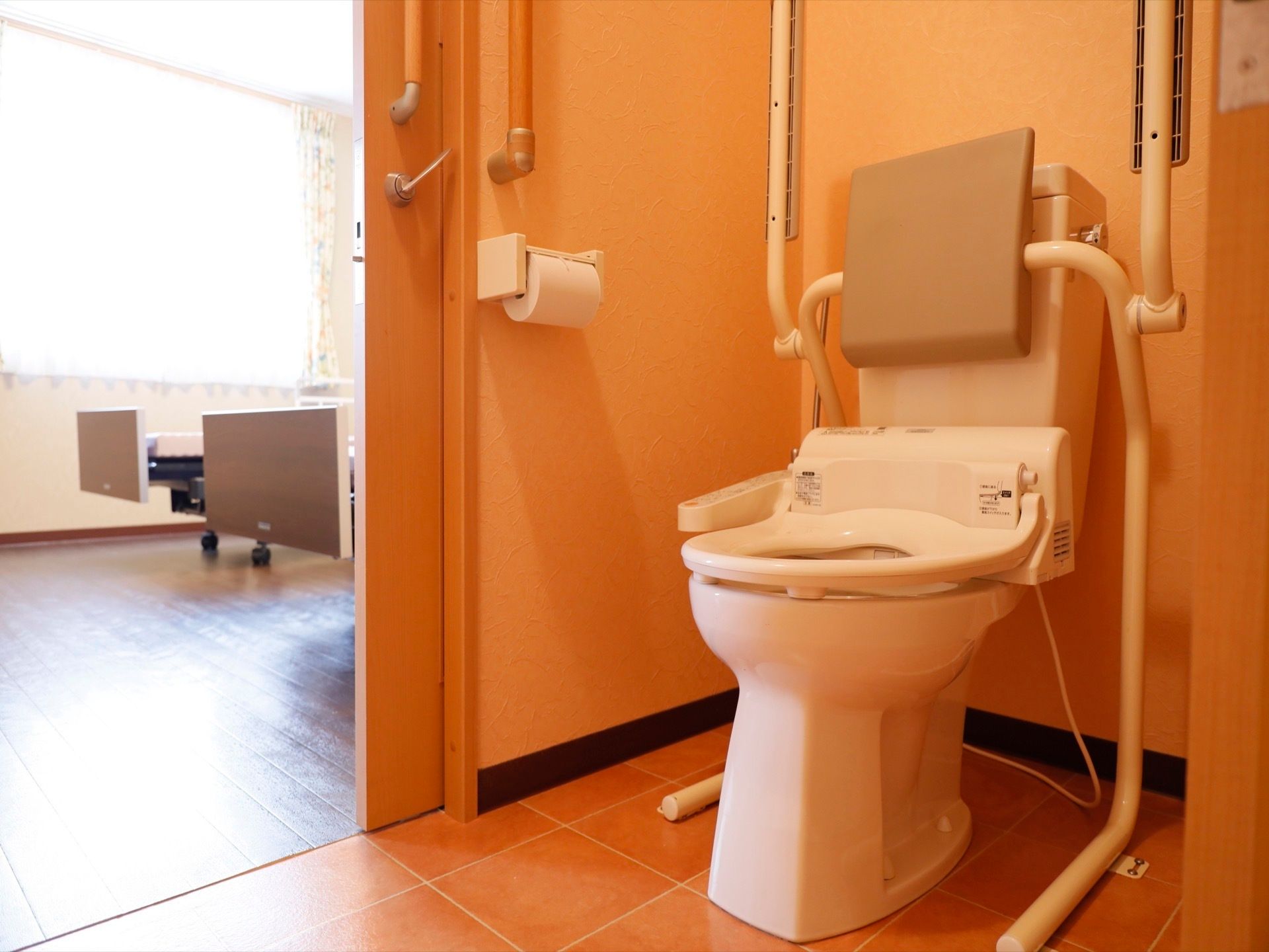 居室のトイレの様子。温水洗浄便座が設置されている。壁の手すりの他に便座に背もたれと可動式の手すりが付いている。
