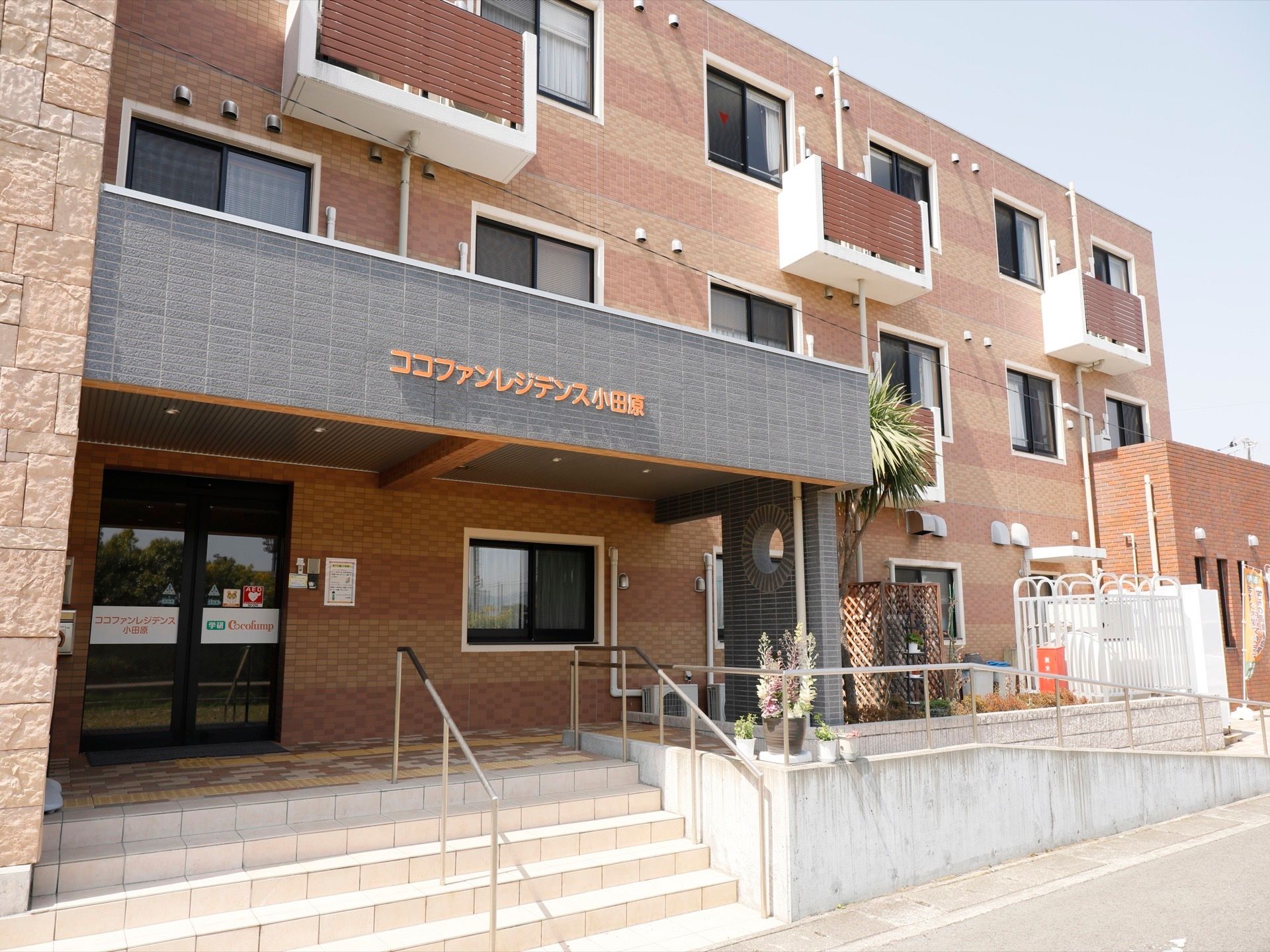 施設外観の様子。レンガ調の３階建ての建物で、手すり付きの階段と手すり付きのスロープが玄関までのびている。