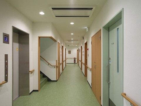 廊下の壁沿いに、入居者様の居室へと続く扉が並んでいる。両側の壁には手すりが取り付けられており、廊下の手前にエレベーターがある。