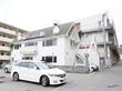 白い２階建ての建物が隣りにあり、屋根の部分に看板がある。行き先案内看板の矢印の方向に進むと、奥に３階建ての大きな施設がある。