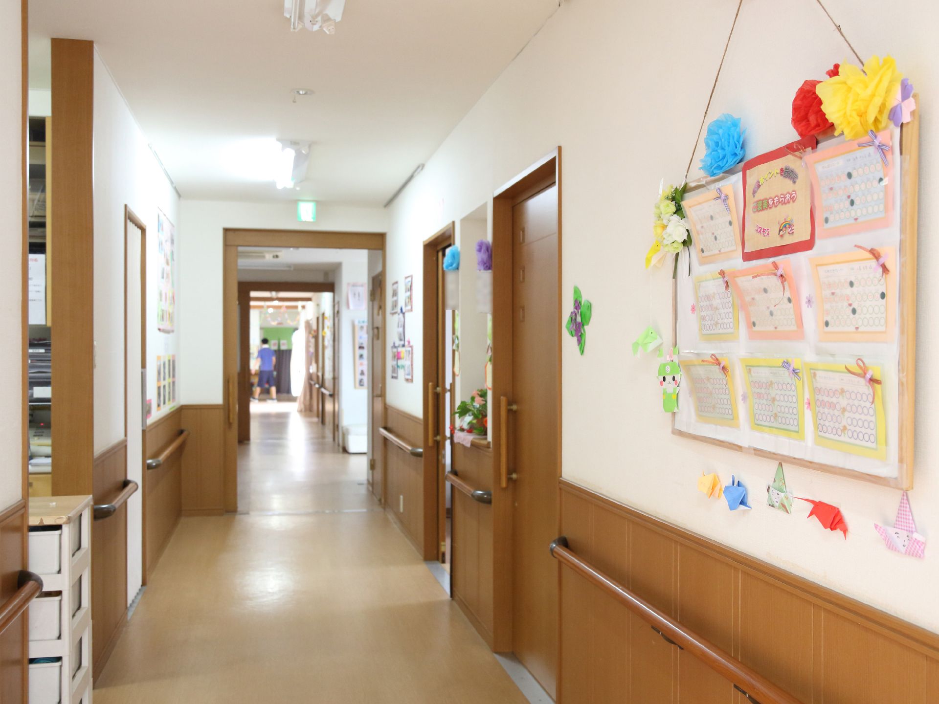 廊下の右側の壁には掲示用のパネルが掛かっており、周りには花や折り紙などが飾り付けられている。床はフローリングである。