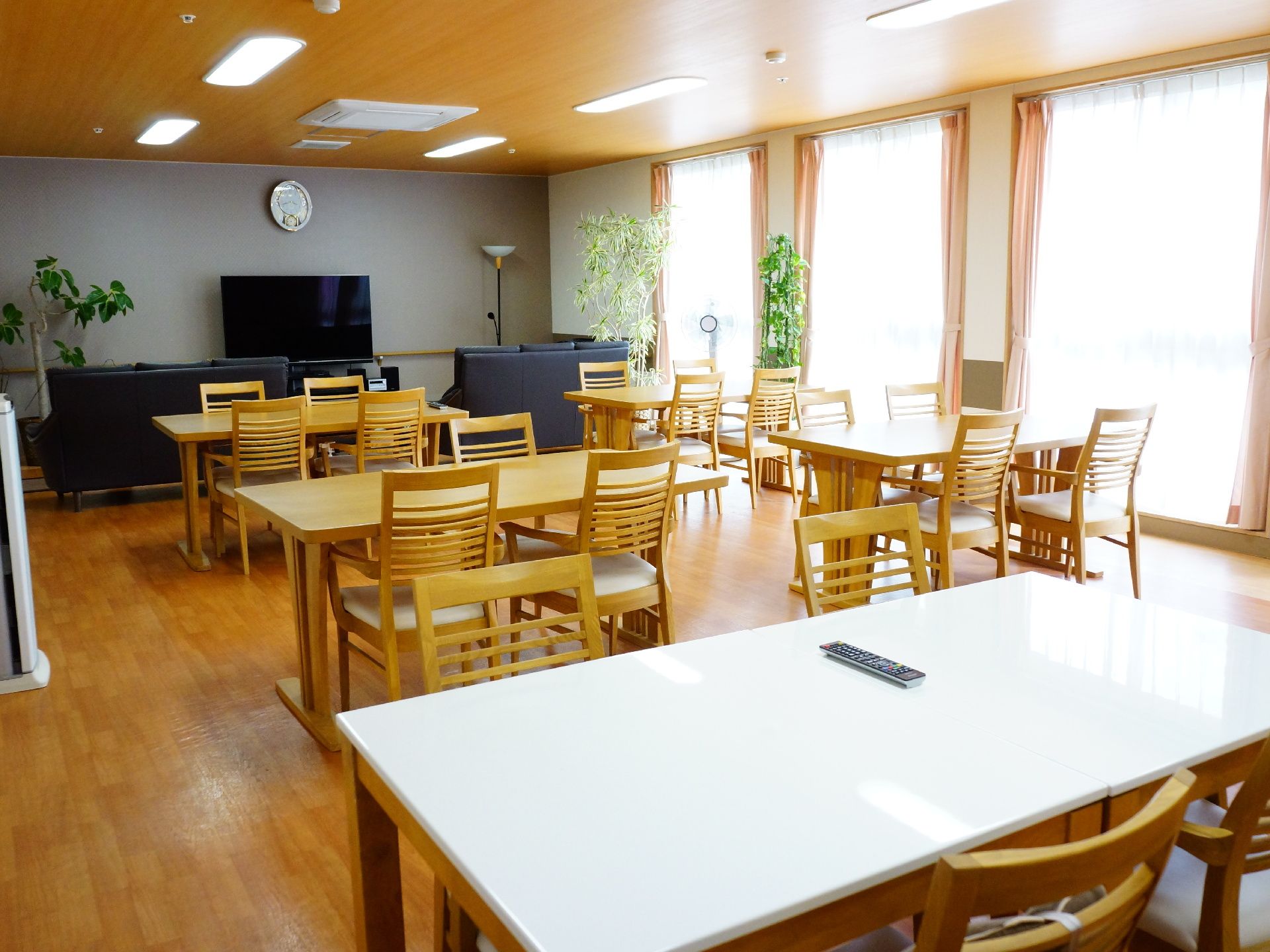 広いフローリングの食堂には大きなガラス戸が並び、明るい日差しが差し込む。奥にはテレビがあり、手前には食事をするテーブルが並んでいる。