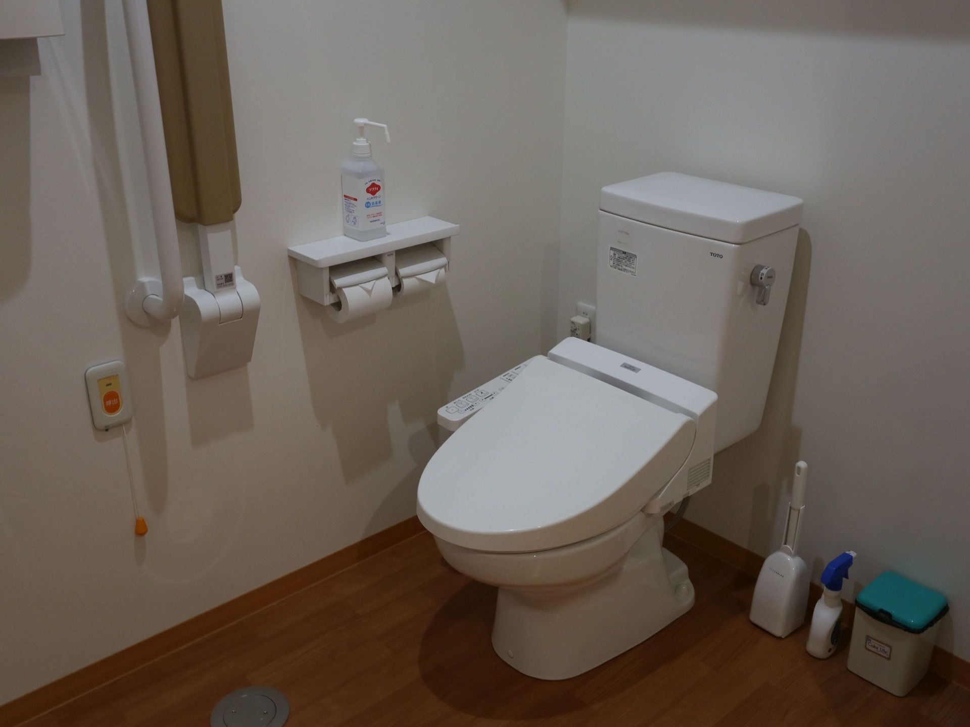 トイレットペーパーホルダーの上には消毒液が置かれている。掃除用具がある。低めの位置に呼び出しボタンがある。