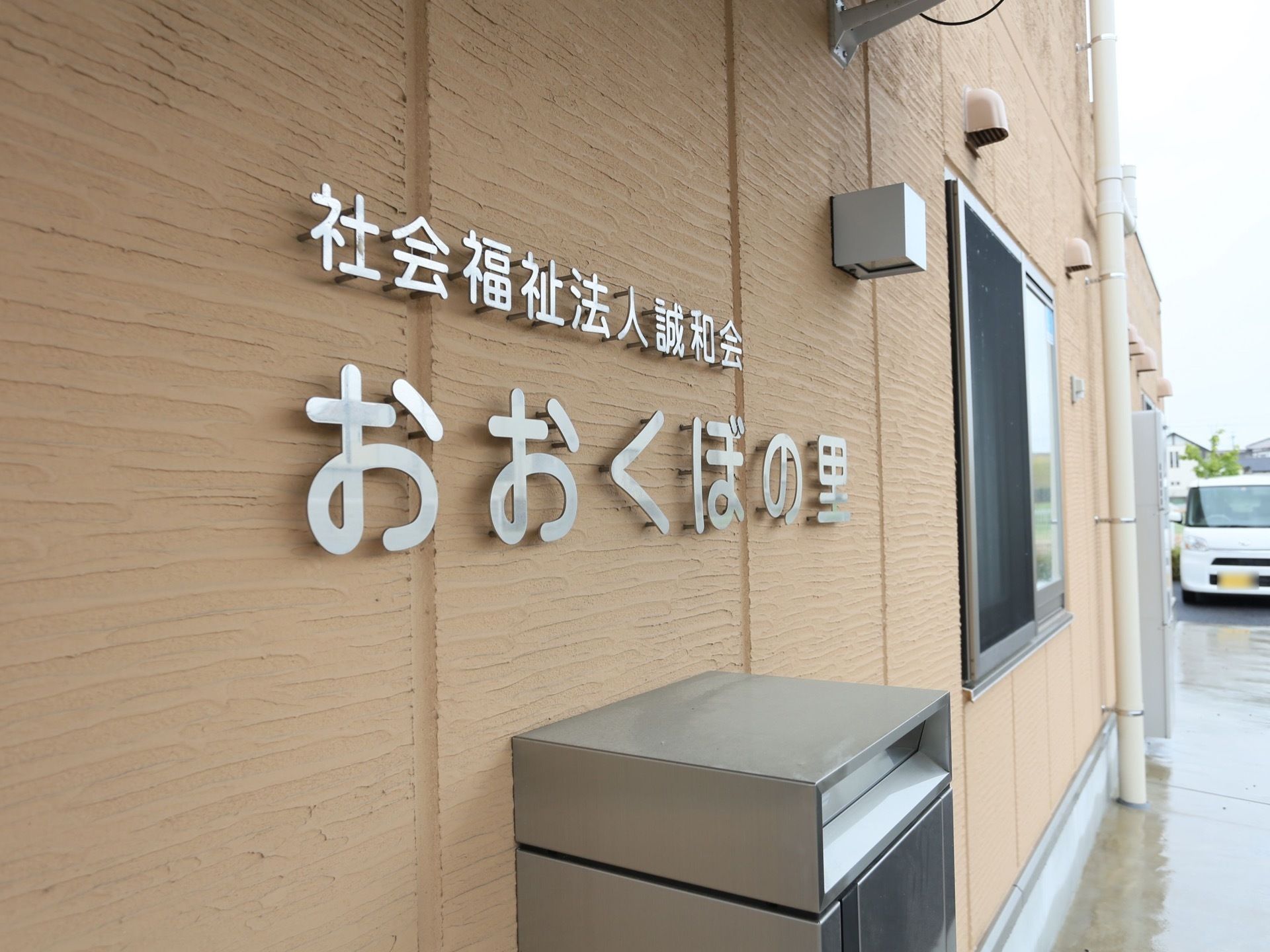 駐車場に車が停まっている。建物は濃いベージュ色で、大きな文字で施設名が書かれている。下には郵便受けが設置されている。