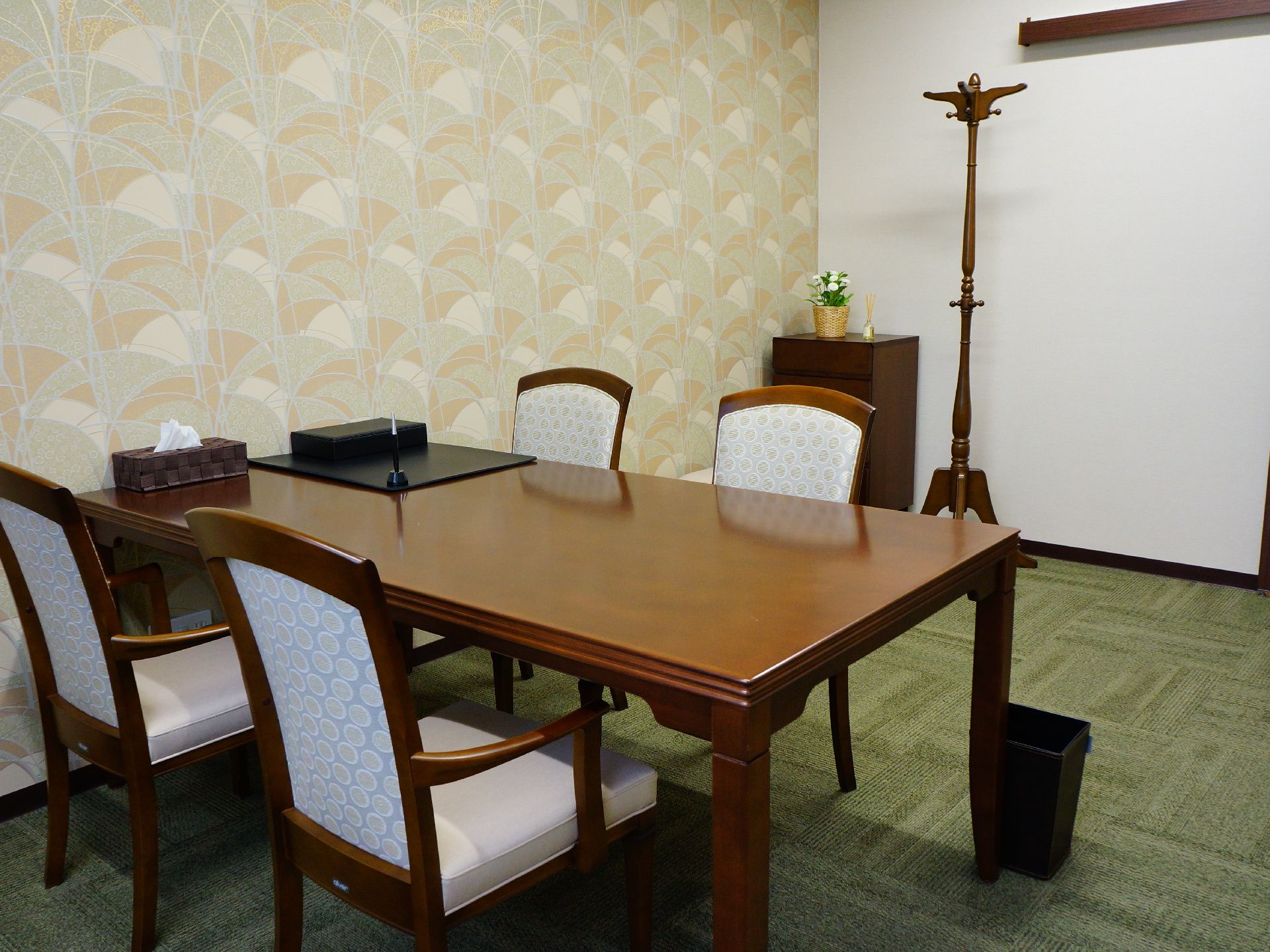 柄の入った壁紙が貼られた部屋に、テーブルセット、コート掛け、棚が置かれている。棚の上には花が置かれている。