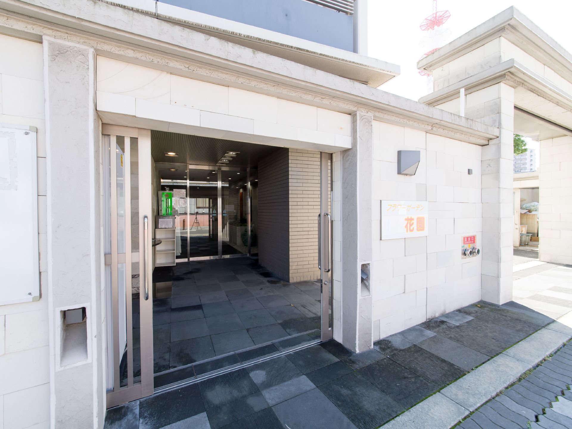 壁の厚い門構えで、看板が取り付けられている。色の違うタイル石が敷かれている。奥には透明の自動ドアがある。