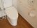 フローリングの床にタンク付きの洋式トイレが設置されている。壁に手すりとペーパーホルダーと温水洗浄便座のリモコンが取り付けられている。