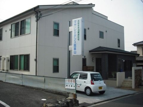 右奥に玄関ポーチが見える、一般家屋のような佇まいの施設である。手前には施設名の書かれた看板があり、その下に白い車が停まっている。