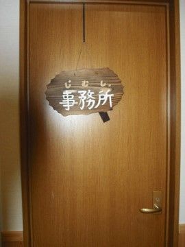 木目調の左開きドアの写真である。事務室と書かれた木製の札がかかっている。周囲の壁は上半分が白、下半分は木の壁になっている。