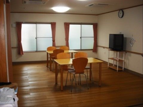 木のフローリングの部屋に角のない机と椅子が置かれており、壁には手すりが取り付けられている。テレビも設置されており、自由に過ごすことができる。