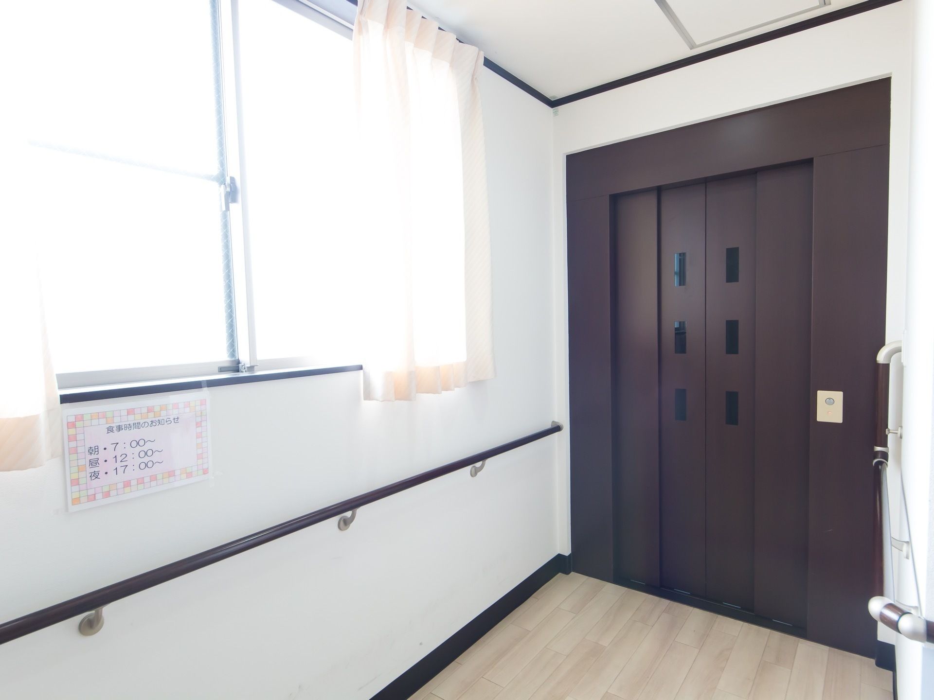 ダークブラウン色の扉のエレベーターが設置されている。そばには窓があり、ベージュ色のカーテンが掛けられている。