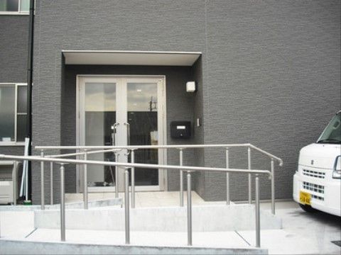 ダークグレーの外観、白い枠にガラスがはめ込まれた扉がある。扉の前は緩やかなスロープで、手すりが取り付けられている。