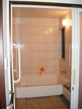 浴室の壁は白い大きな壁のタイルが貼られている。浴室側の壁には白い横長の手すりがある。手すりにはオレンジ色の手桶が置かれている。
