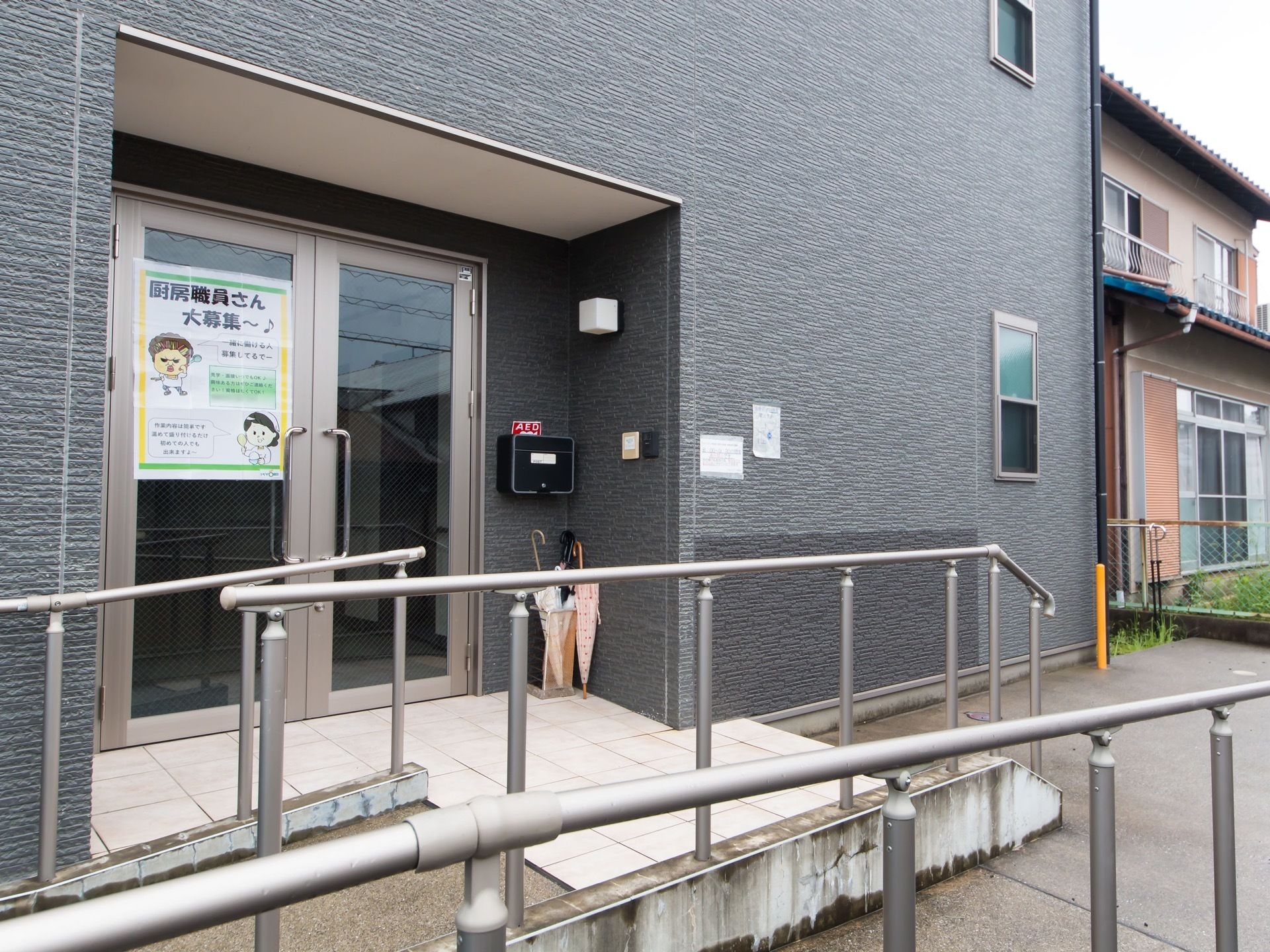 舗装された敷地に建てられた濃い灰色の施設である。玄関の前方にスロープや階段、手すりがある。