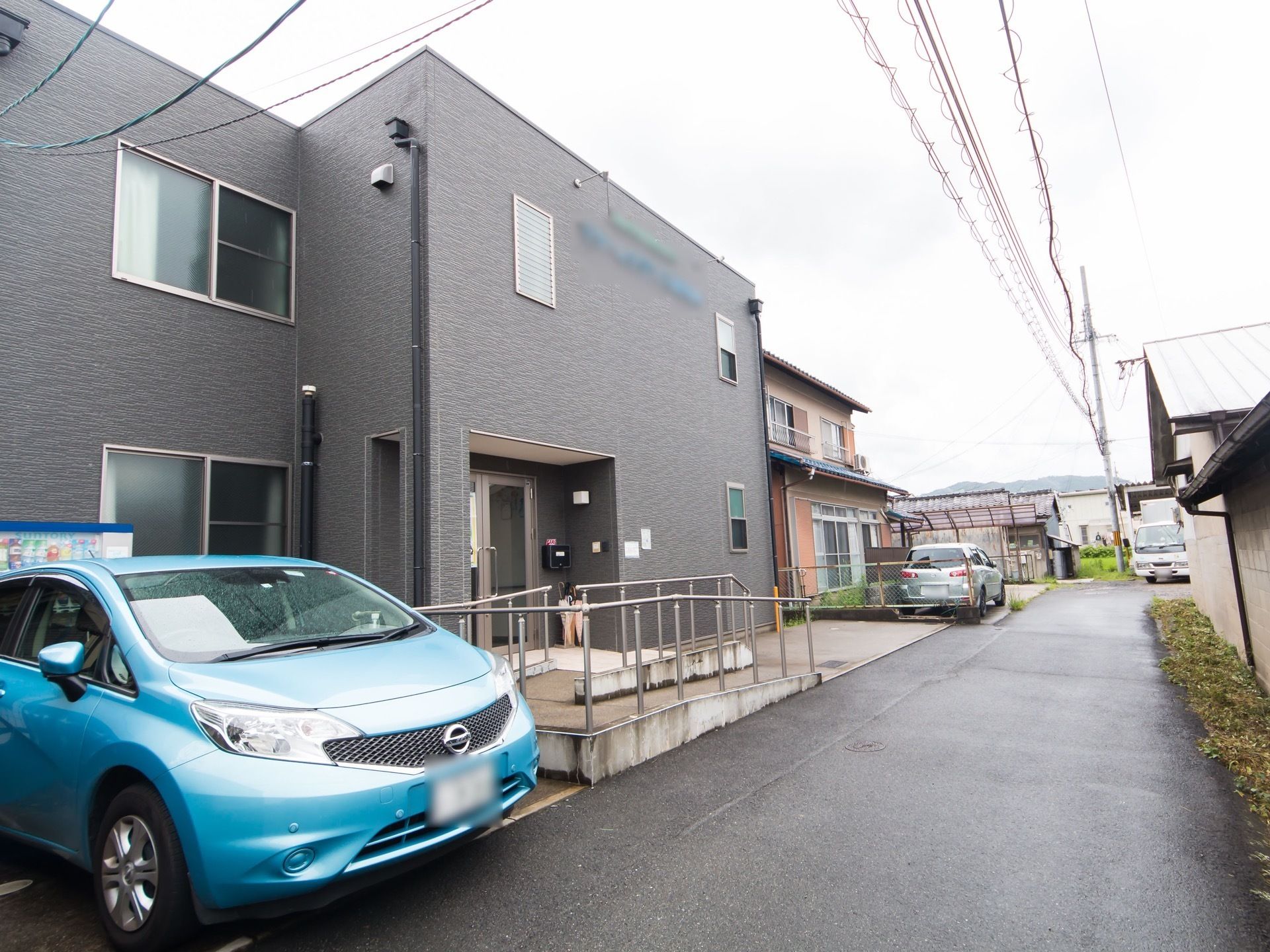 住宅街の一角に２階建ての施設が建っており、手前に青い自動車が停めてある。右側の奧には田畑や山が見える。