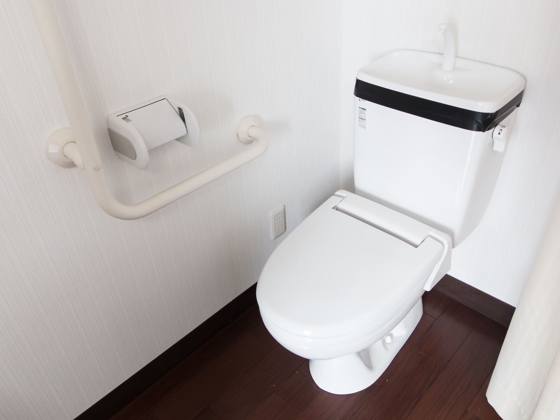 タンクや蓋の付いた洋式便器が据えてあるトイレスペースである。左側の壁面にはＬ字型の手すりがある。