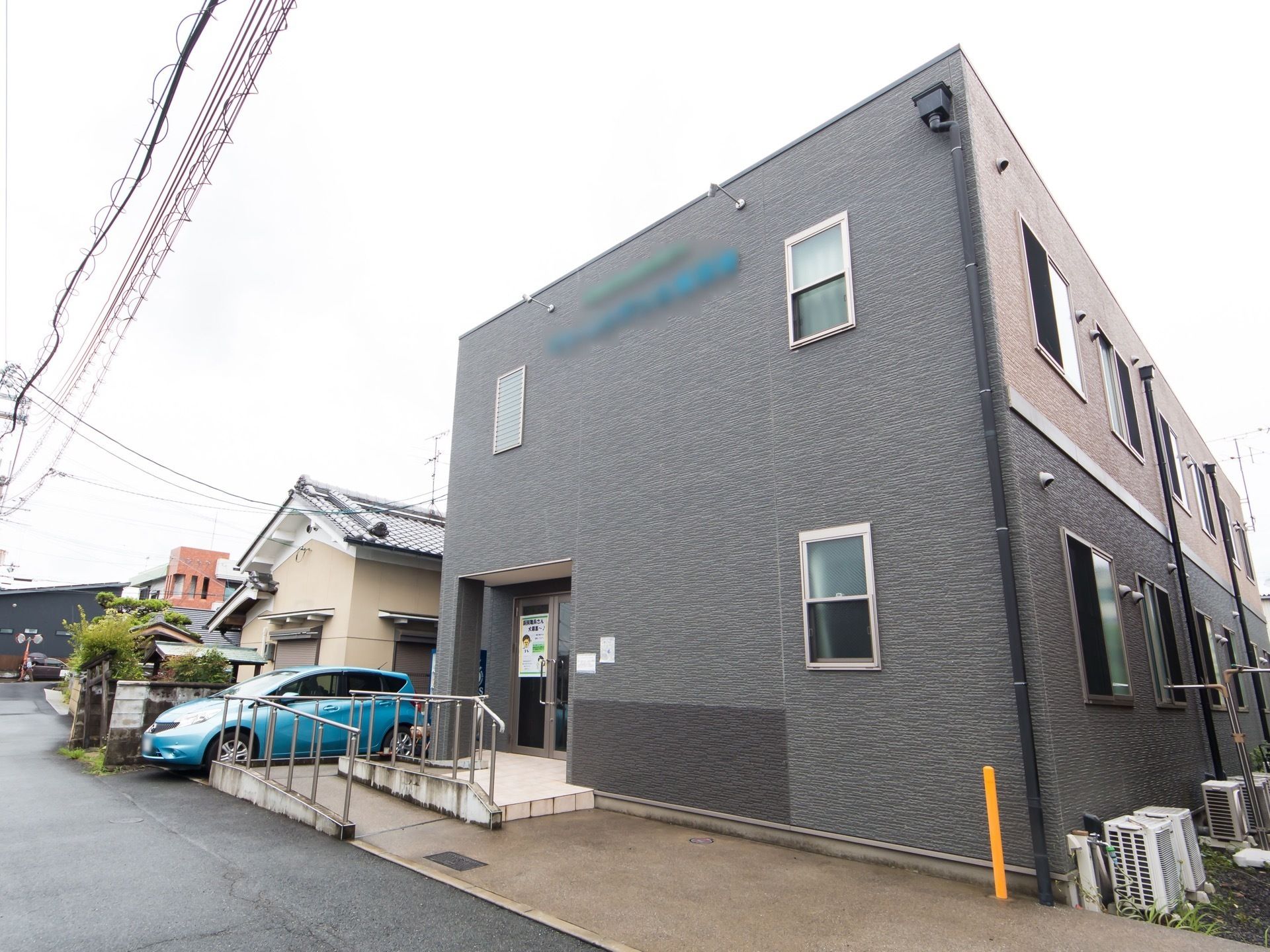 濃い灰色の外壁が施された２階建ての施設が道路に面して建っている。建物の前に自動車が１台停められている。