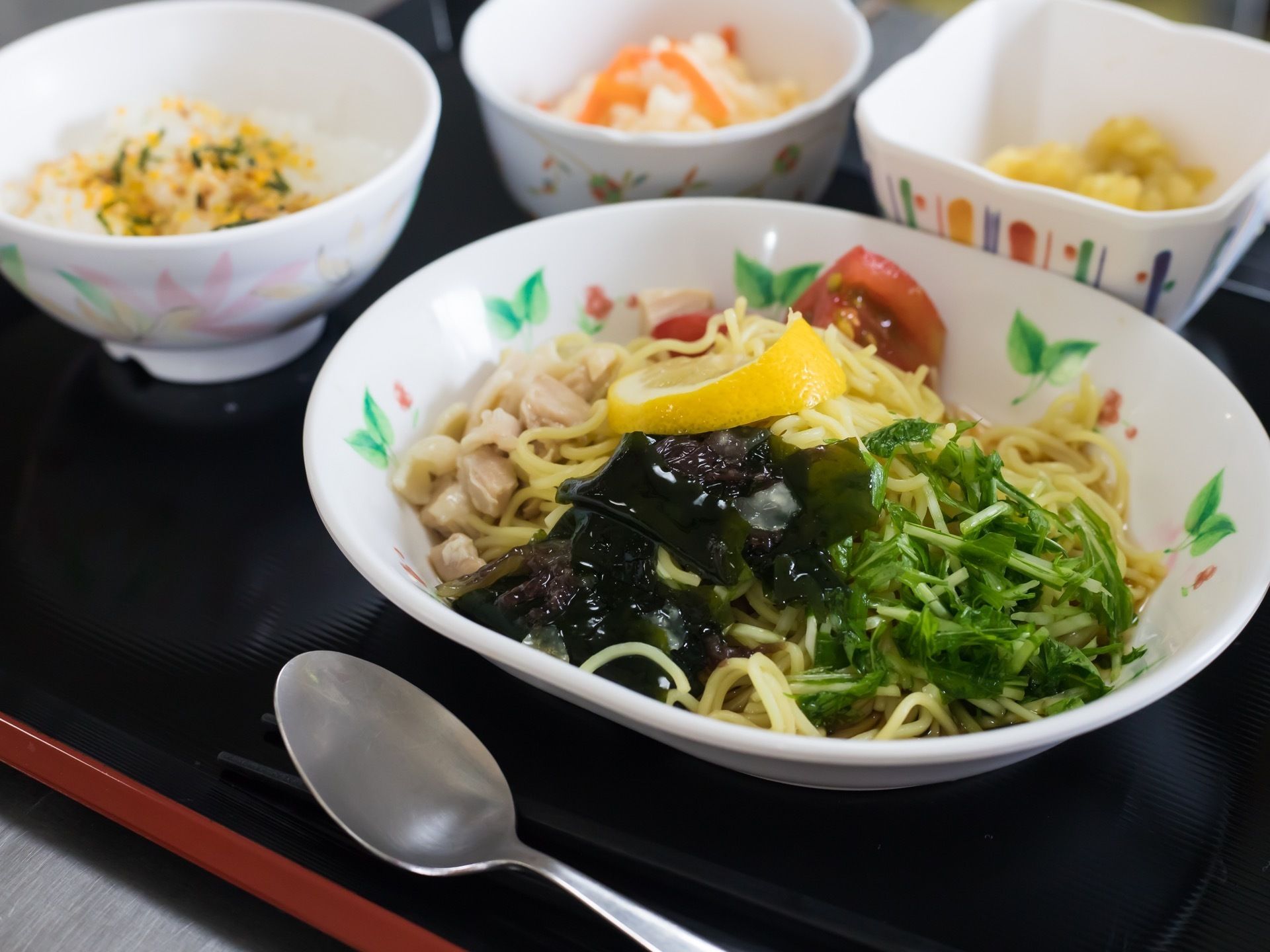 黒いトレイの上に複数の小鉢、箸とスプーンが用意されている。ふりかけが掛かったごはん、麺類、２種類のおかずが見える。