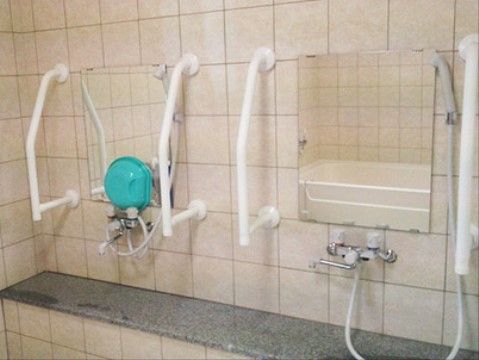 鏡が横に２枚並んでいる浴室。鏡の両横に手すりがある。すぐ下にある蛇口の片方には、水色の洗面器がのせられている。