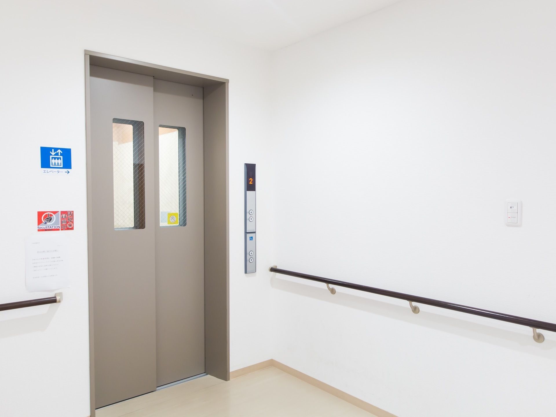通路の突き当たりに設置されたエレベーター。低い位置に操作ボタンがある。白い壁には黒い手すりが伸びている。