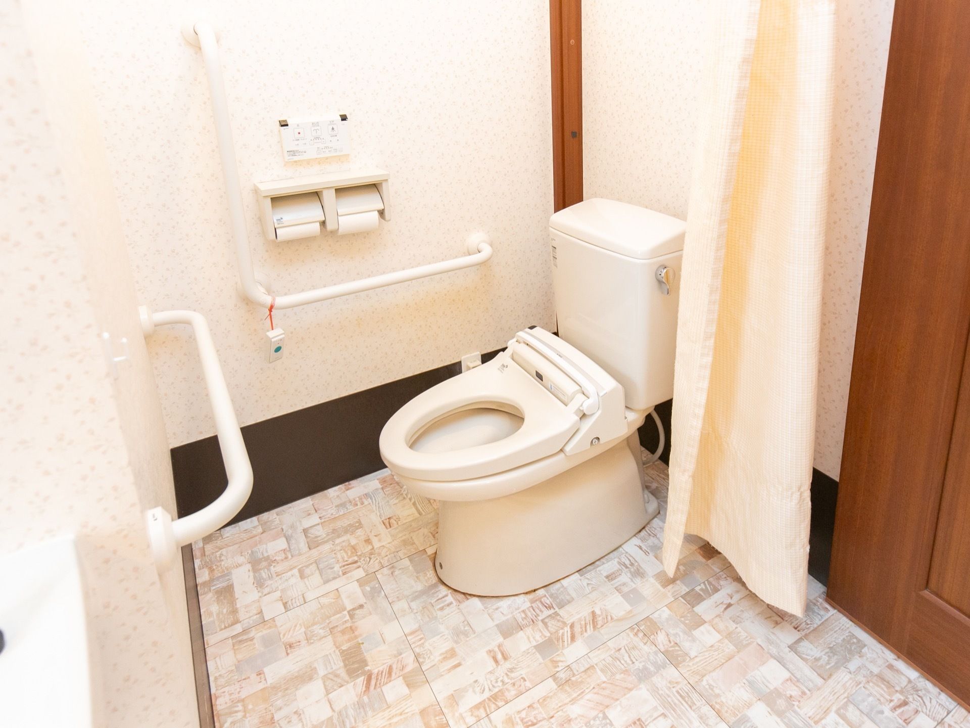 壁には手すり、ペーパーホルダー、緊急呼び出しボタンが設置されている。目隠しカーテンが付けられている。