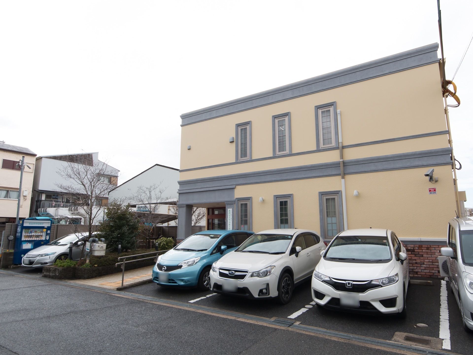 建物の前方には駐車スペースがあり、車が複数止まっている。玄関アプローチには植え込みが造られていて、木が植えられている。