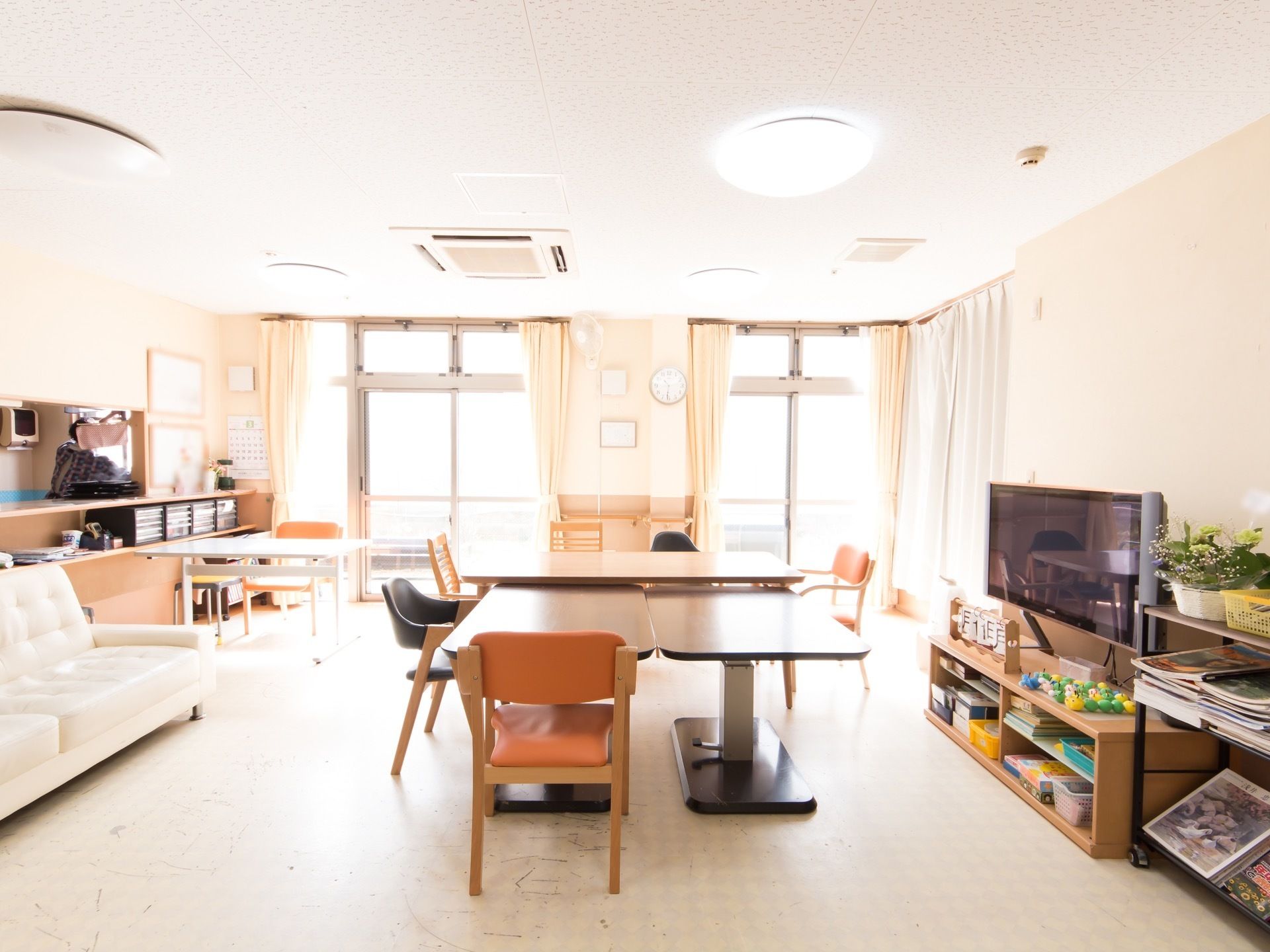 食堂には大きな窓が設けられていて、白いカーテンが掛かっている。手前の壁際にはテレビ台と大画面のテレビが設置されている。