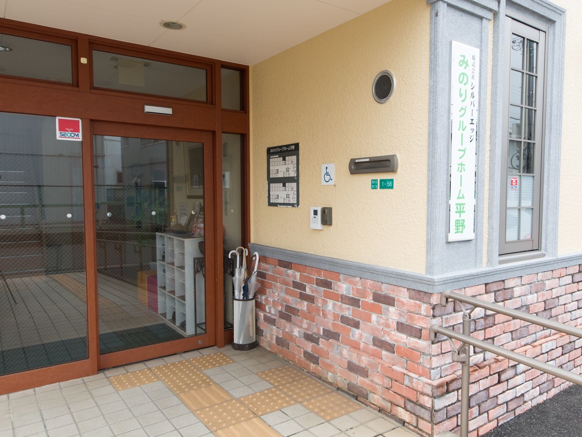 レンガが埋められた玄関ポーチで、壁にポストと館内案内図が貼られている。床に円柱形の傘立てが置かれている。