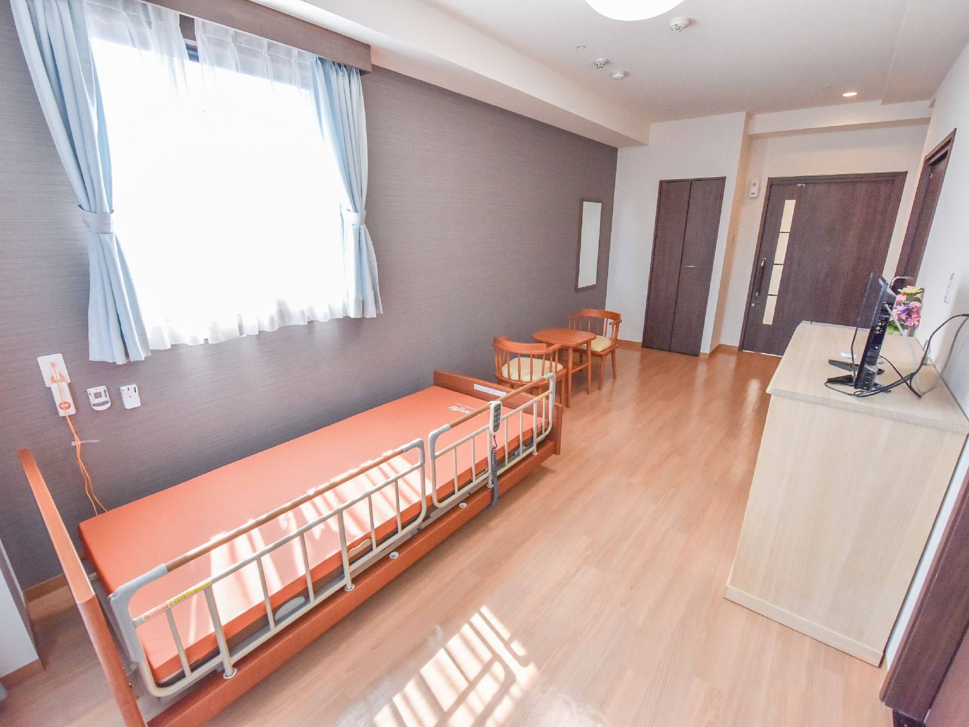 ウッドフロアで、白い壁の部屋で、布団の落下防止の柵がついた電動ベッドがあるほか、テーブルと椅子がある。
