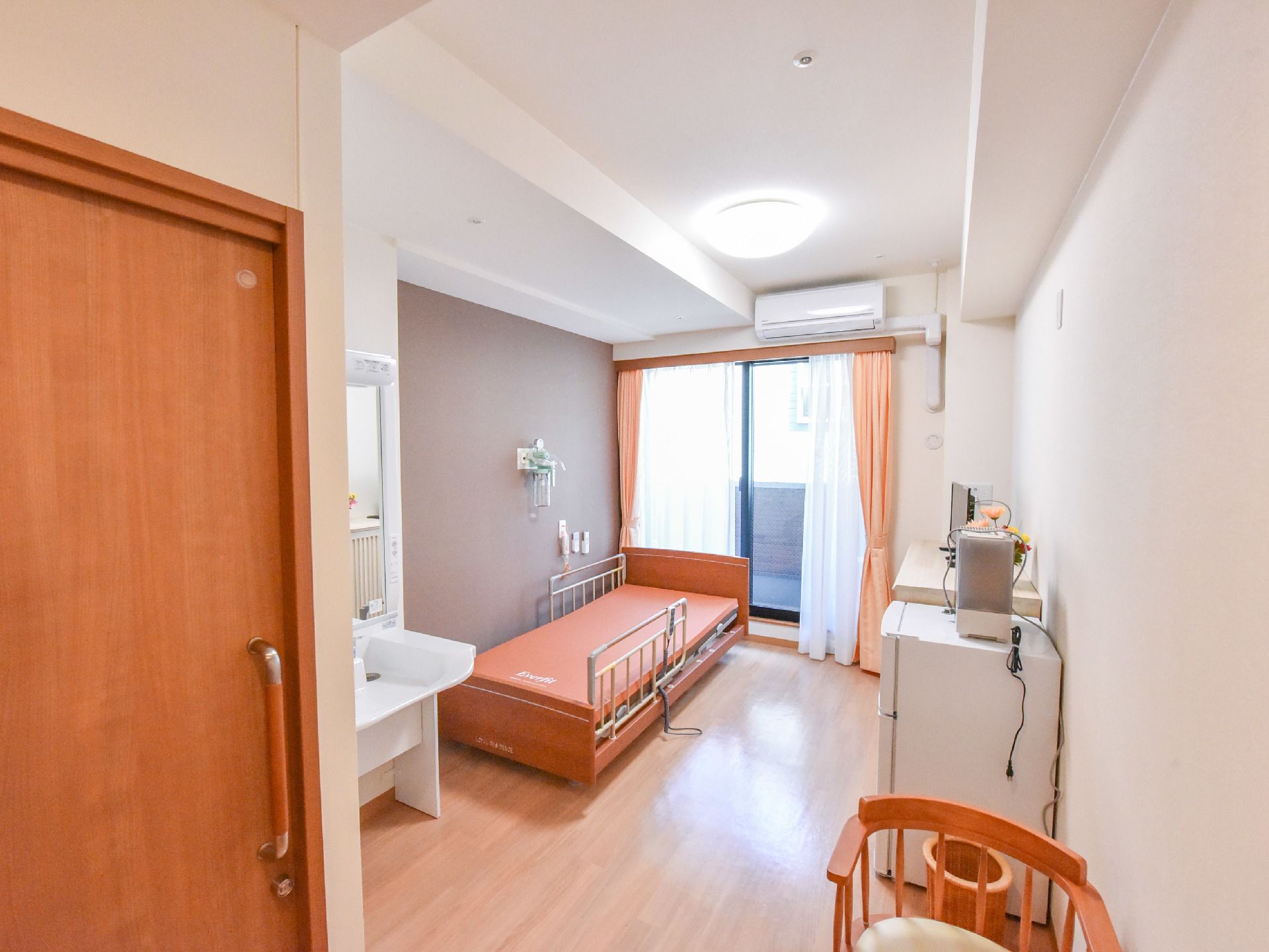 白い壁と天井、床は茶色の部屋に、白い洗面台が置いてある。茶色の電動ベッドには落下防止の柵が取り付けてある。