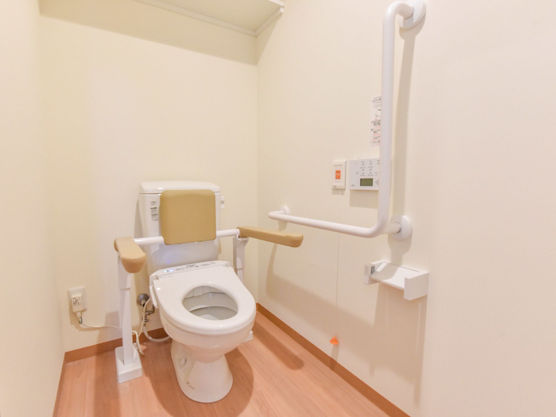 フローリングの床でできているトイレで、両サイドに手すり、背あてがついている。壁に緊急時用のブザーが設置してある。