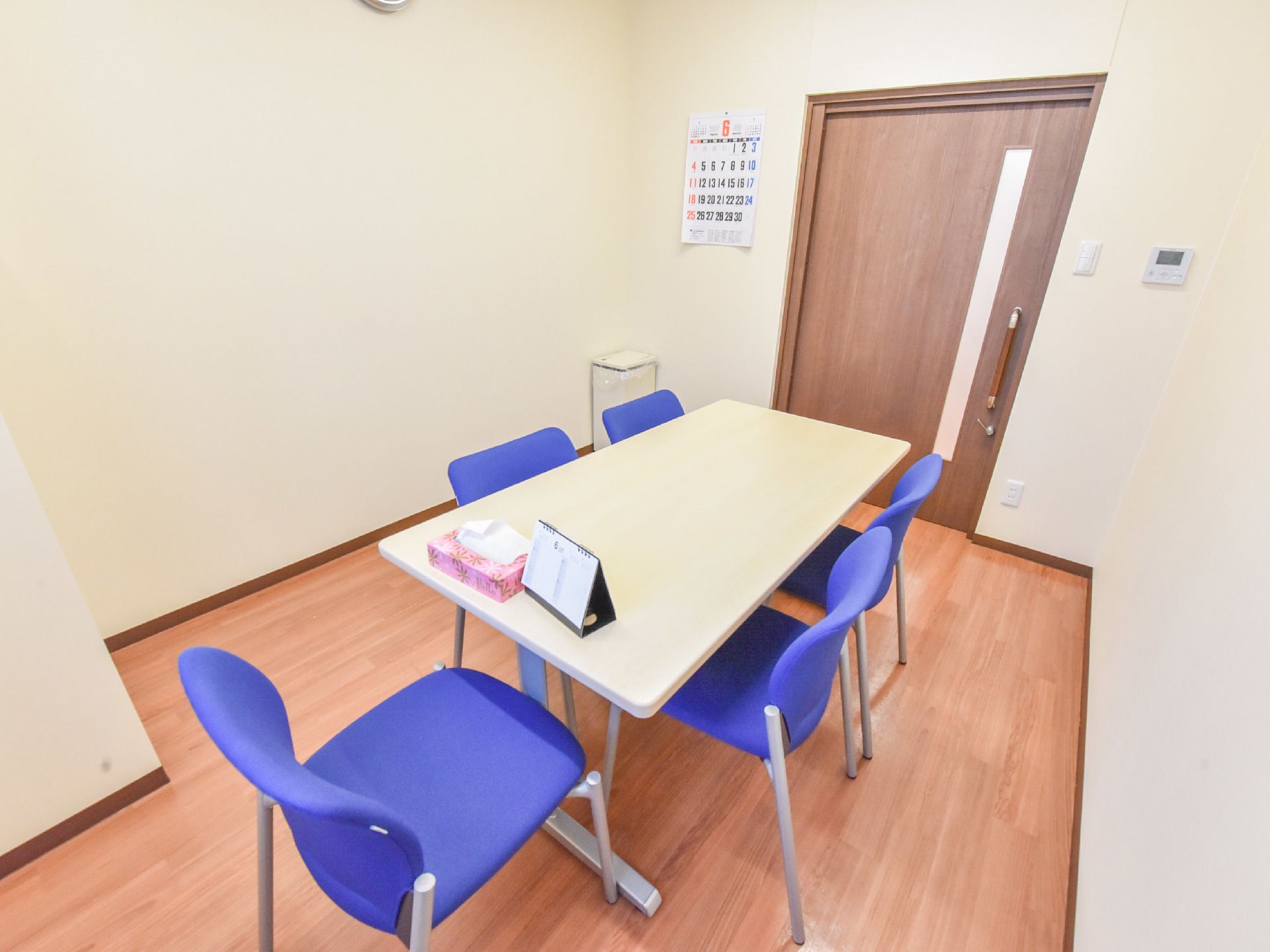 白いテーブルと青い椅子が置いてある部屋で、白い壁で構成されている。扉は茶色である。壁に時計がかけてある。