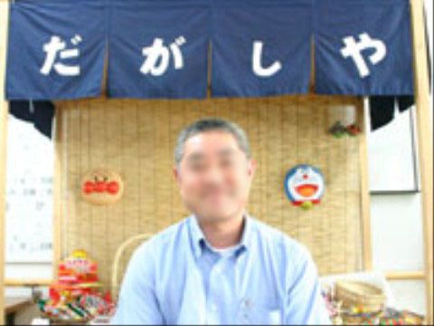 「だがしや」と文字の入った濃紺の暖簾が下がった下に、１人の男性がいる。ザルに入れられた駄菓子やキャラクターのお面が飾ってある。