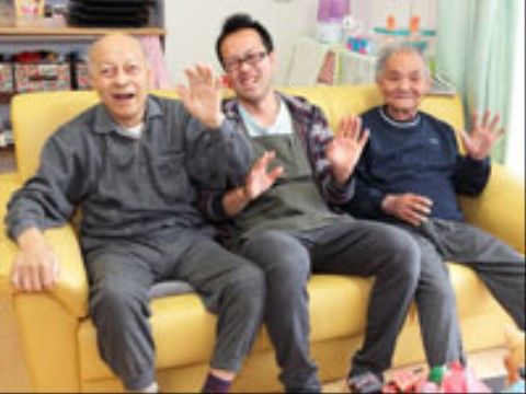 黄色のソファに座って笑顔でポーズをとる２名の入居者と介護スタッフ