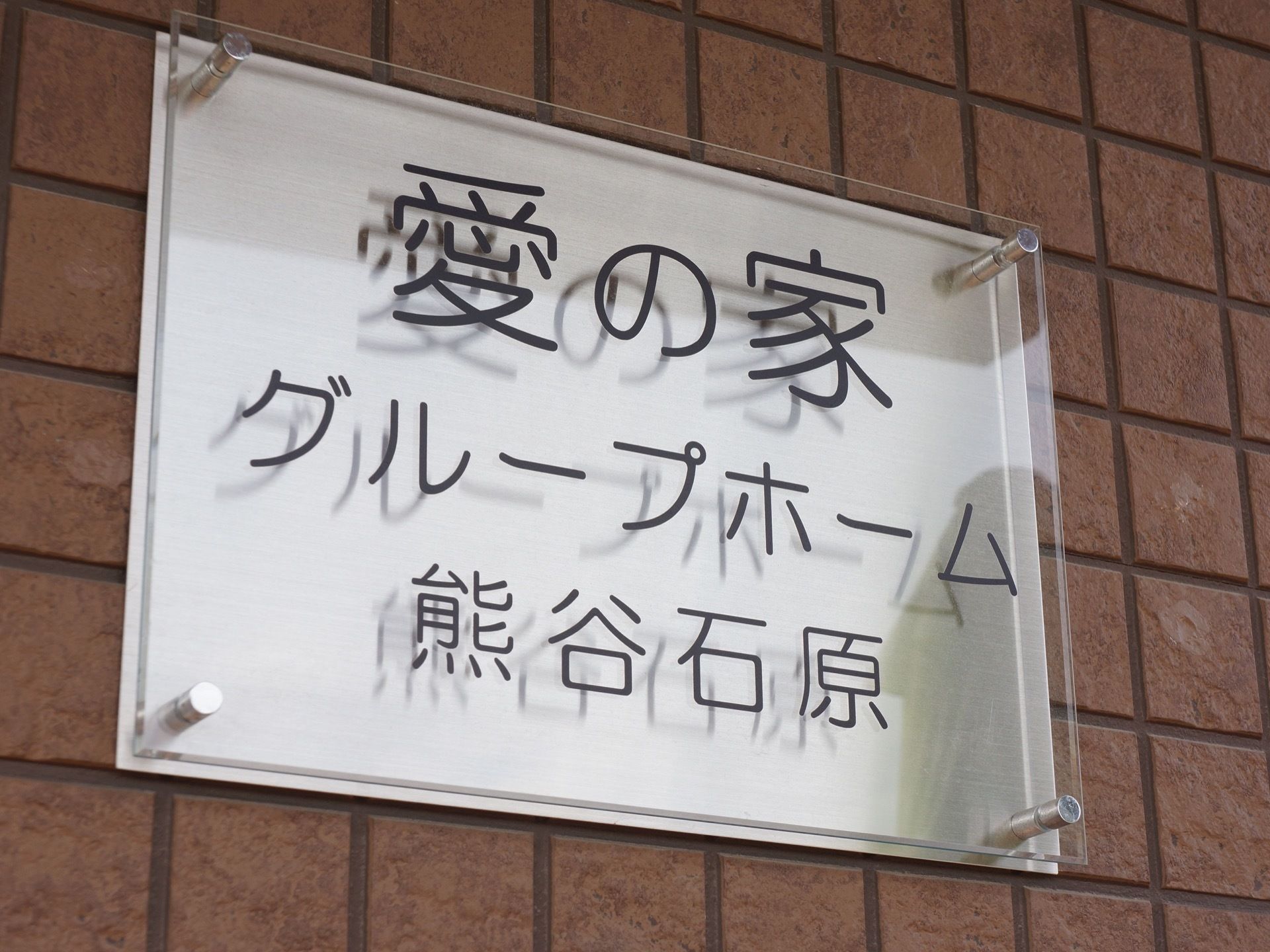 銀色のステンレスのプレートに、アクリルの透明樹脂板がボルトで固定されている。施設表示板で大きな黒文字で見やすい。