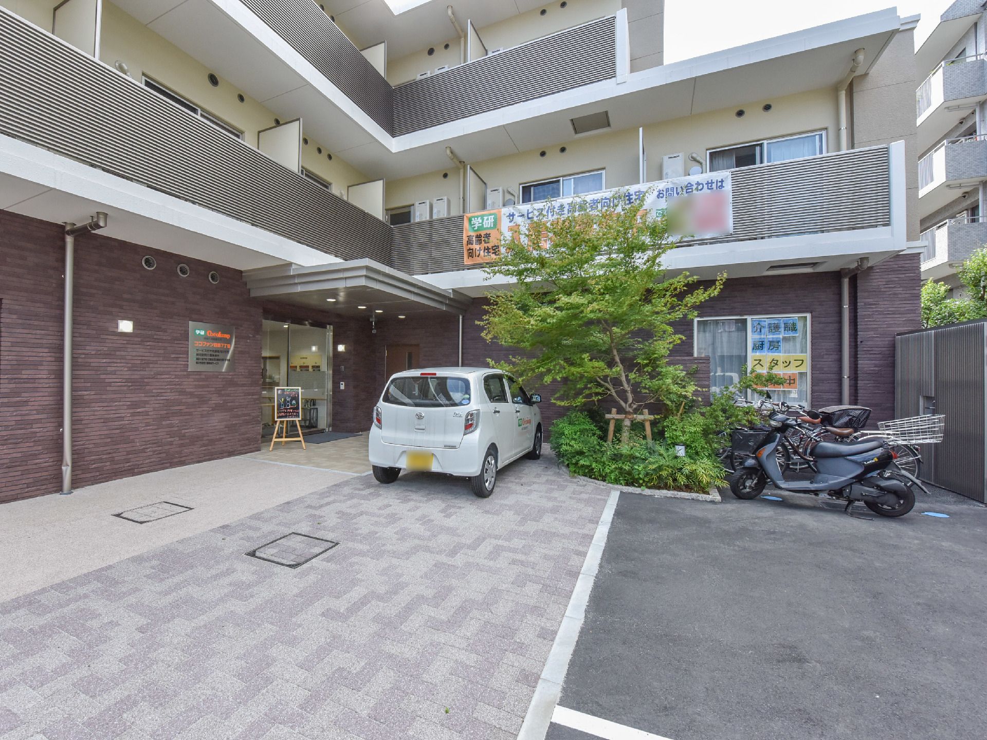 施設の入り口付近の床はベージュ系のタイル敷きである。背の高い木が植えられた植え込みがあり、左側に車が1台停められている。