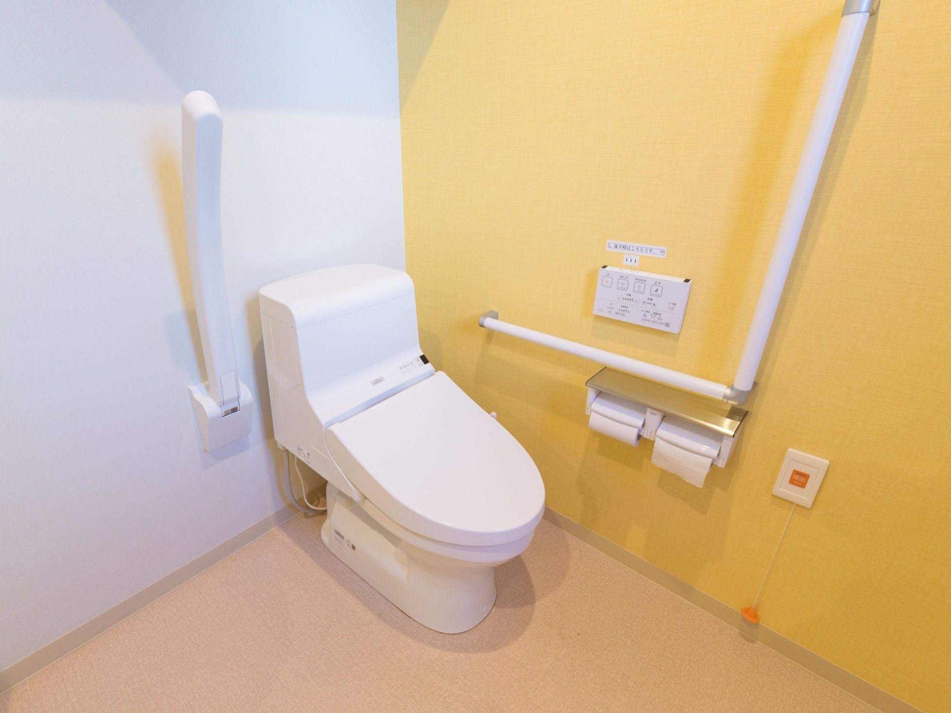 温水洗浄機能付きのトイレの両側には、大きな手すりが付いている。右側の壁には、オレンジ色の緊急用呼出ボタンがある。