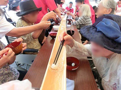 長いテーブルの真ん中に竹の流しそうめんが流れ、向かい合って食べる人々