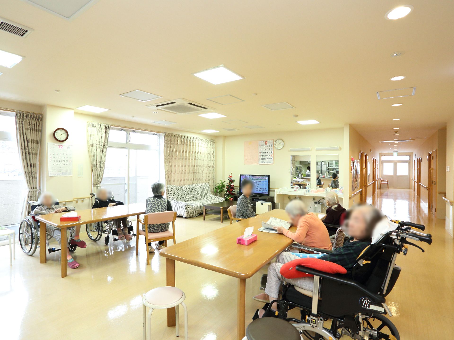 長テーブルが二台置かれ、入居者様が座っている。奥には、テレビモニターやソファも置かれている。手洗い台がテレビの右側にある。