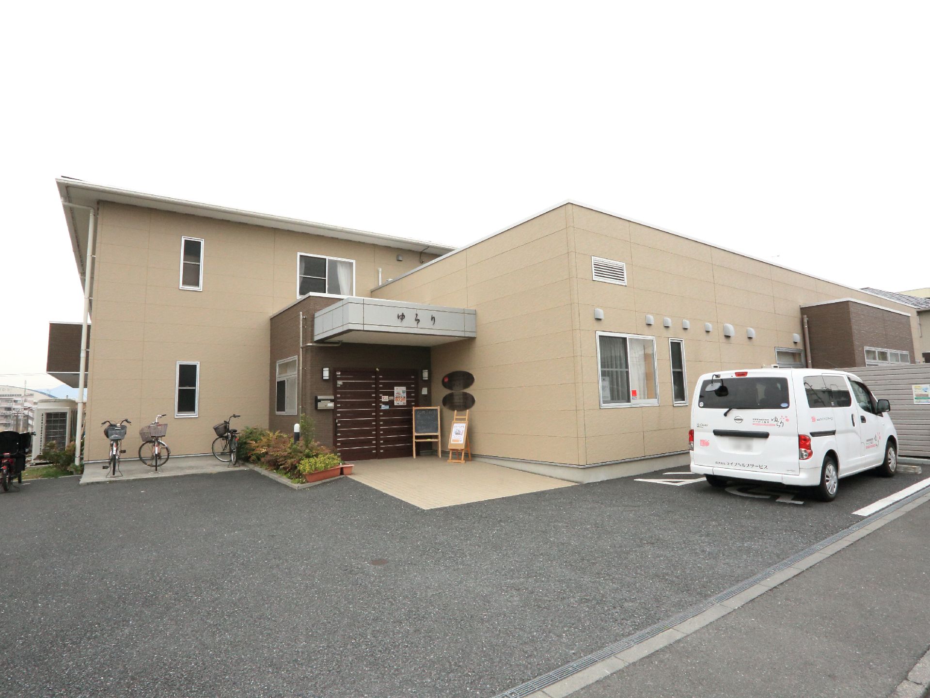 施設外観の様子。玄関前には広くスペースがとられており、右側には一台分の駐車場がある。建物は二階建てとなっている。