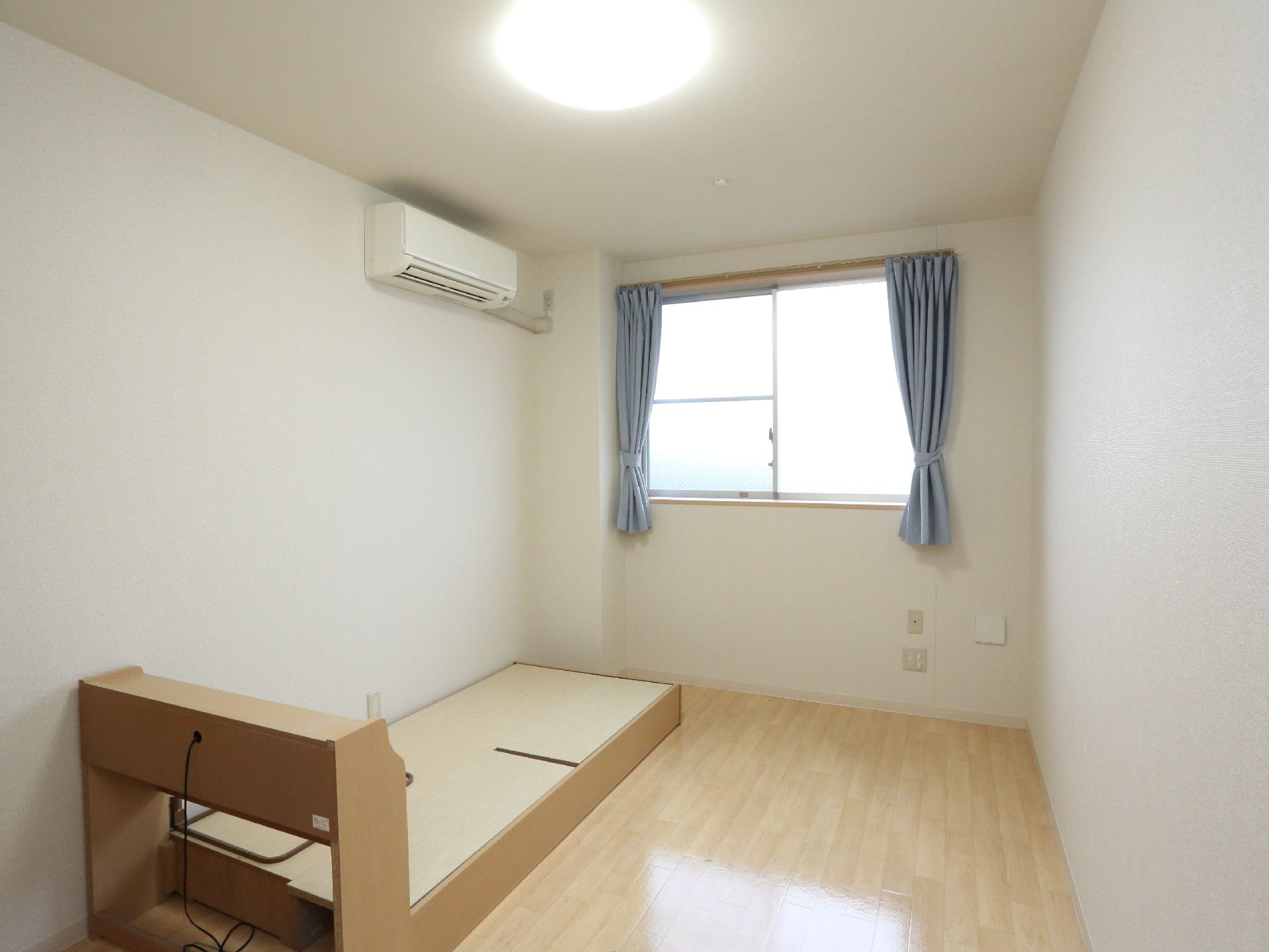 居室の奥に窓があり、カーテンが付いている。壁にエアコンが設置されており、床にベッドが置かれている。床はフローリングである。