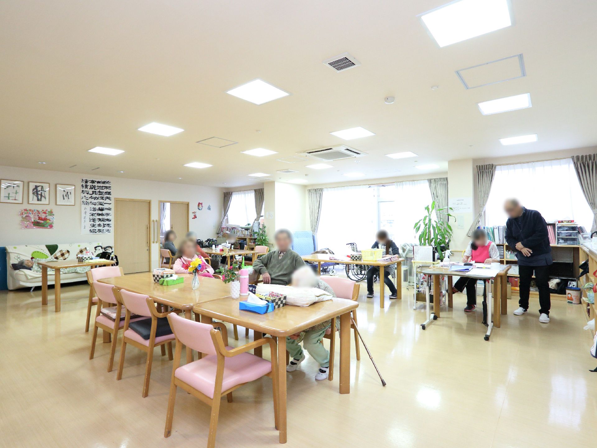 ダイニングは広い空間で、大きな窓から日が差し込んでいる。室内に、入居者様やスタッフがおり、椅子に座ったり作業をしたりしている。
