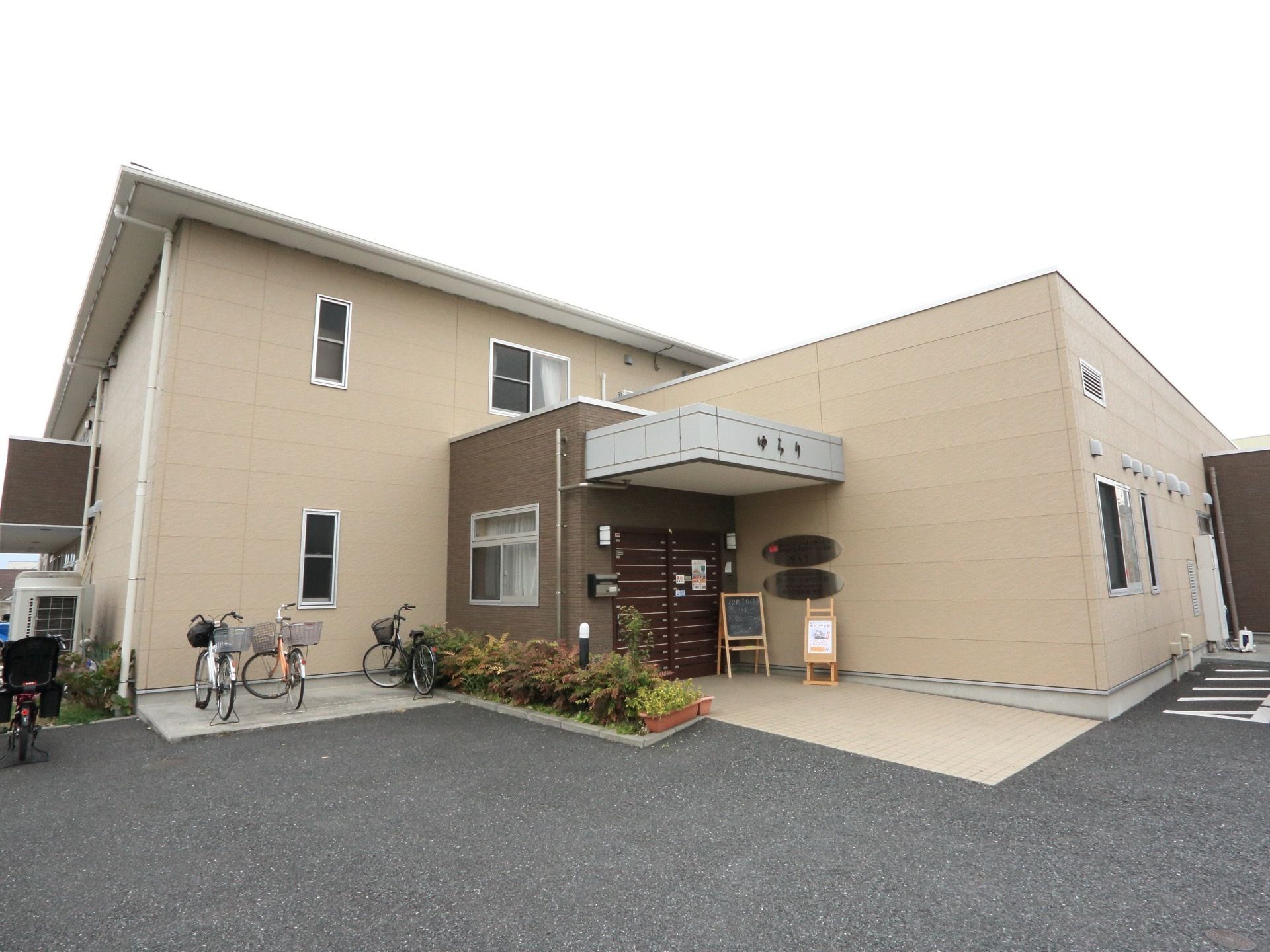駐車場から施設入口まで、段差のない造りになっている。ベージュ色メインの2階建てである。入口周辺に自転車が数台置かれている。