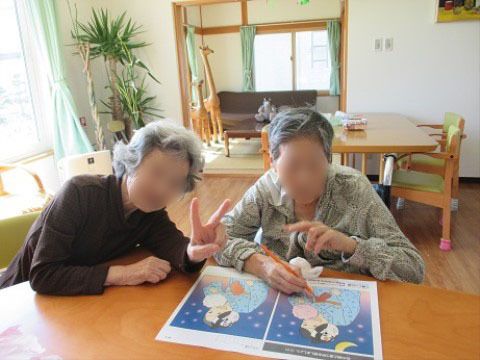 明るい談話室で、ダイニングセットに腰かけ笑顔でポーズをとる入居者様。テーブル上には雑誌が広げられている。