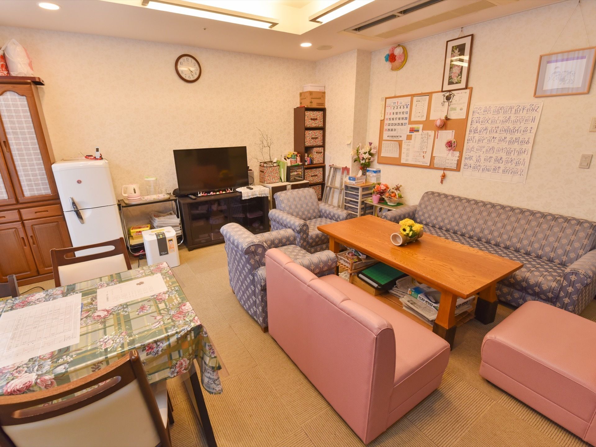 部屋の奥にテレビや冷蔵庫が置かれている。右手には机があり、机を囲むようにソファが置かれている。左手には、テーブルセットがある。